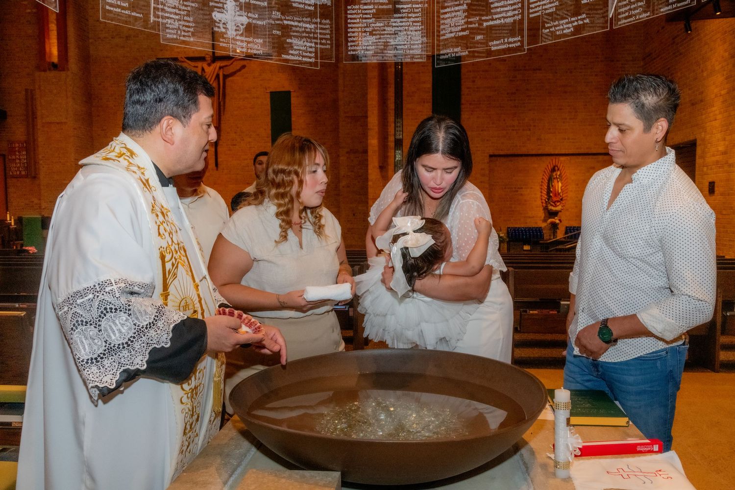Catholic priest performing a baptism ceremony over a font with family members holding an infant in a church.
