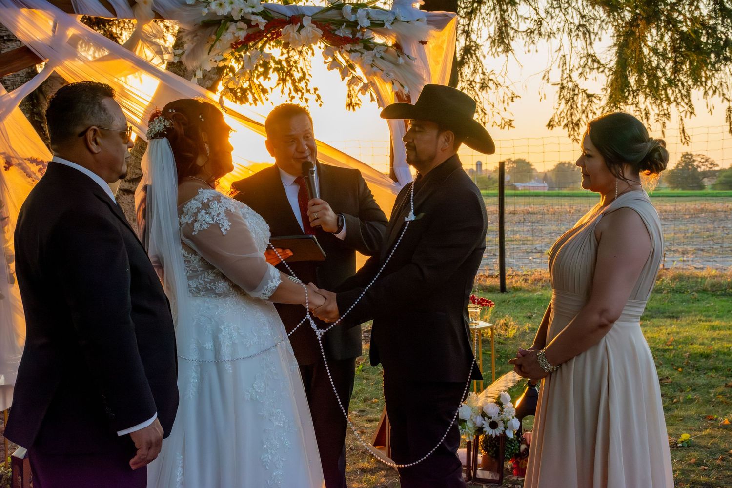 Outdoor wedding ceremony at sunset with couple exchanging vows under a floral arch with officiant.