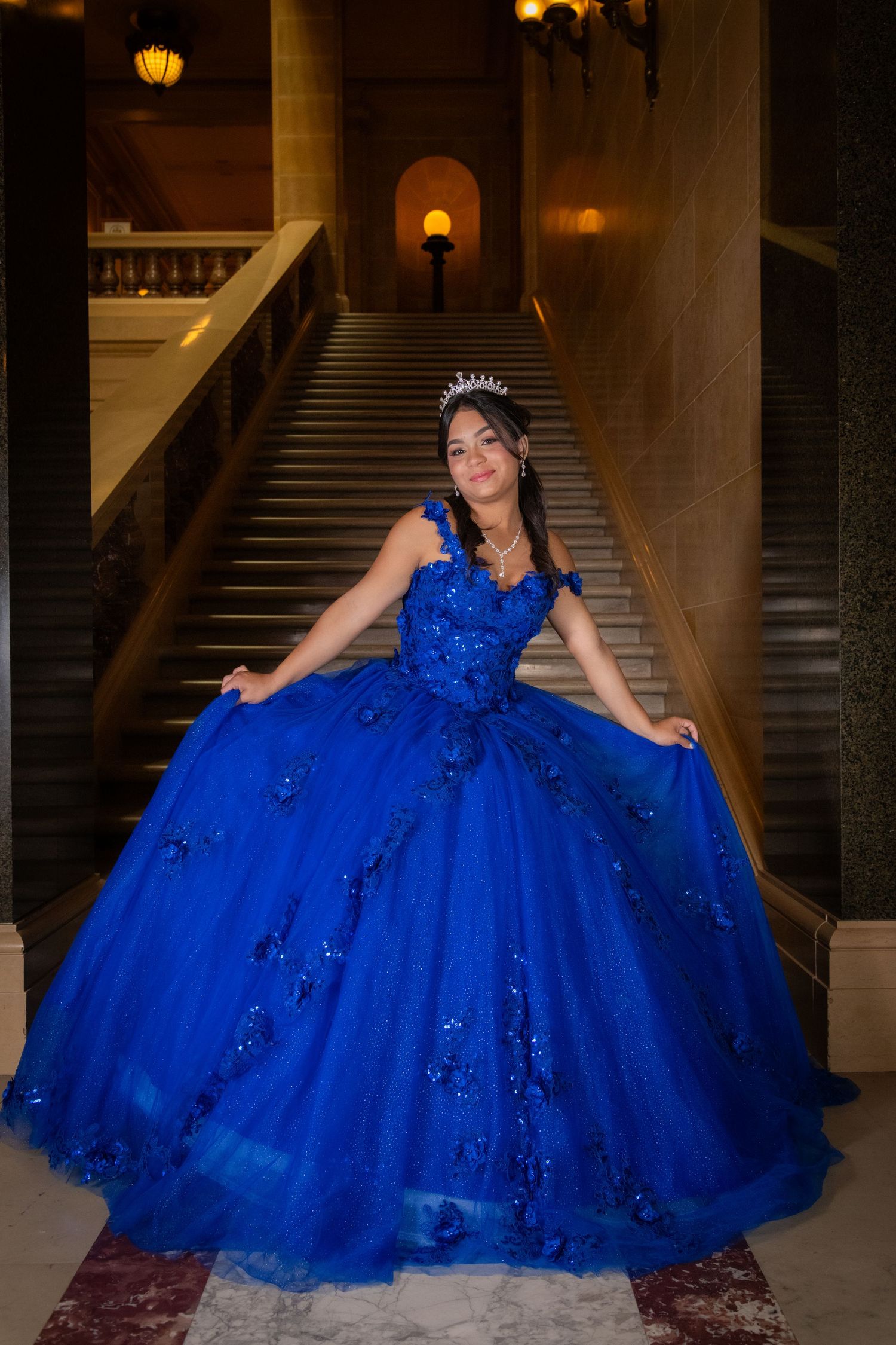 Girl in royal blue ball gown and tiara posing gracefully on grand staircase.