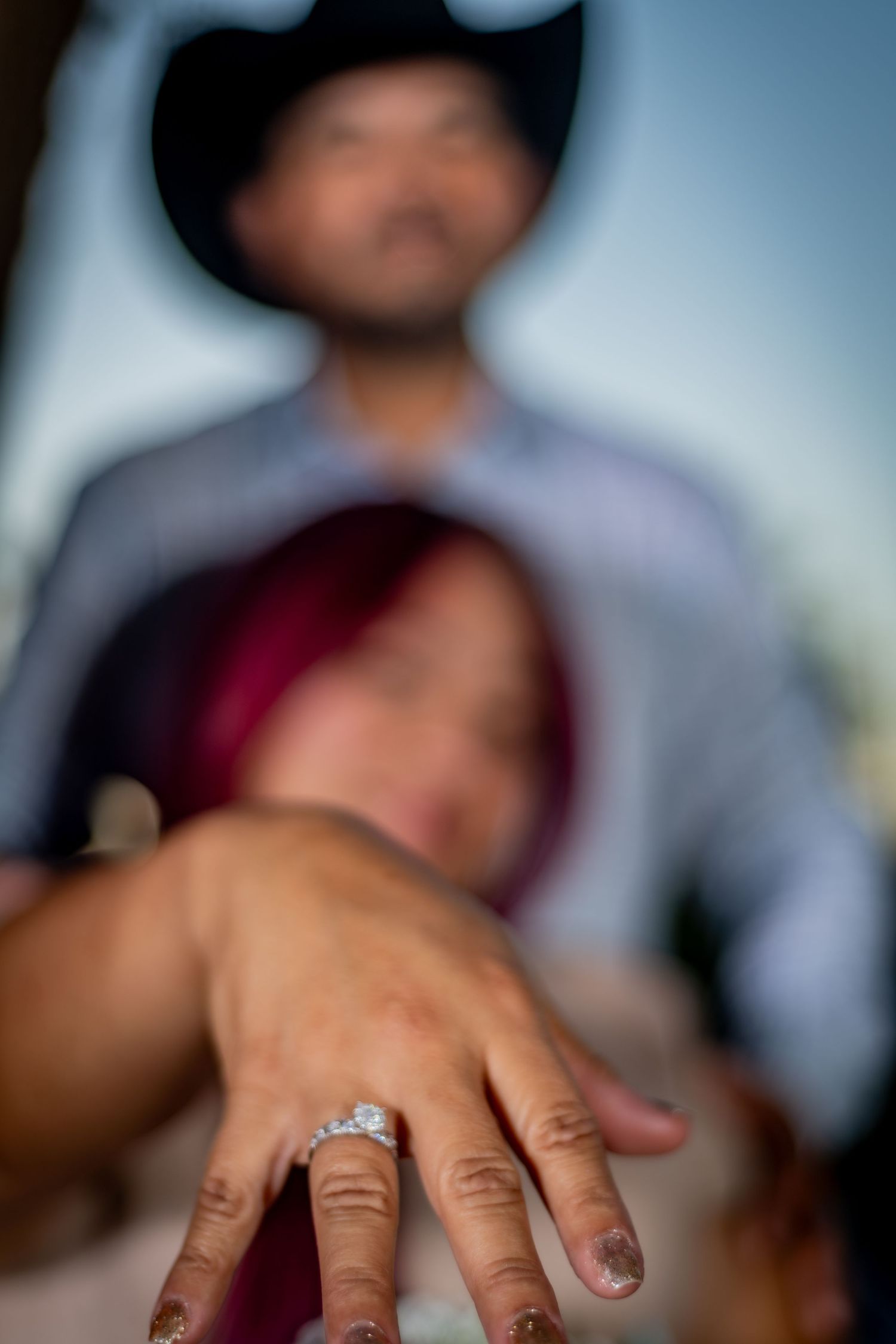 Close-up of an engagement ring on a woman's hand with couple in soft focus background outdoors.