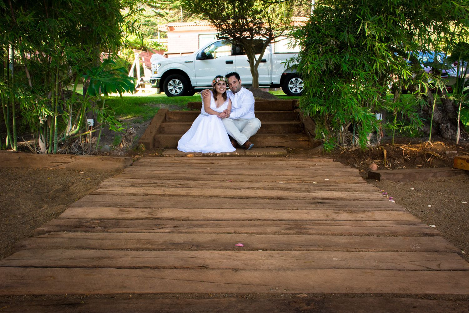Bride and groom sitting together on wooden steps surrounded by lush green trees.