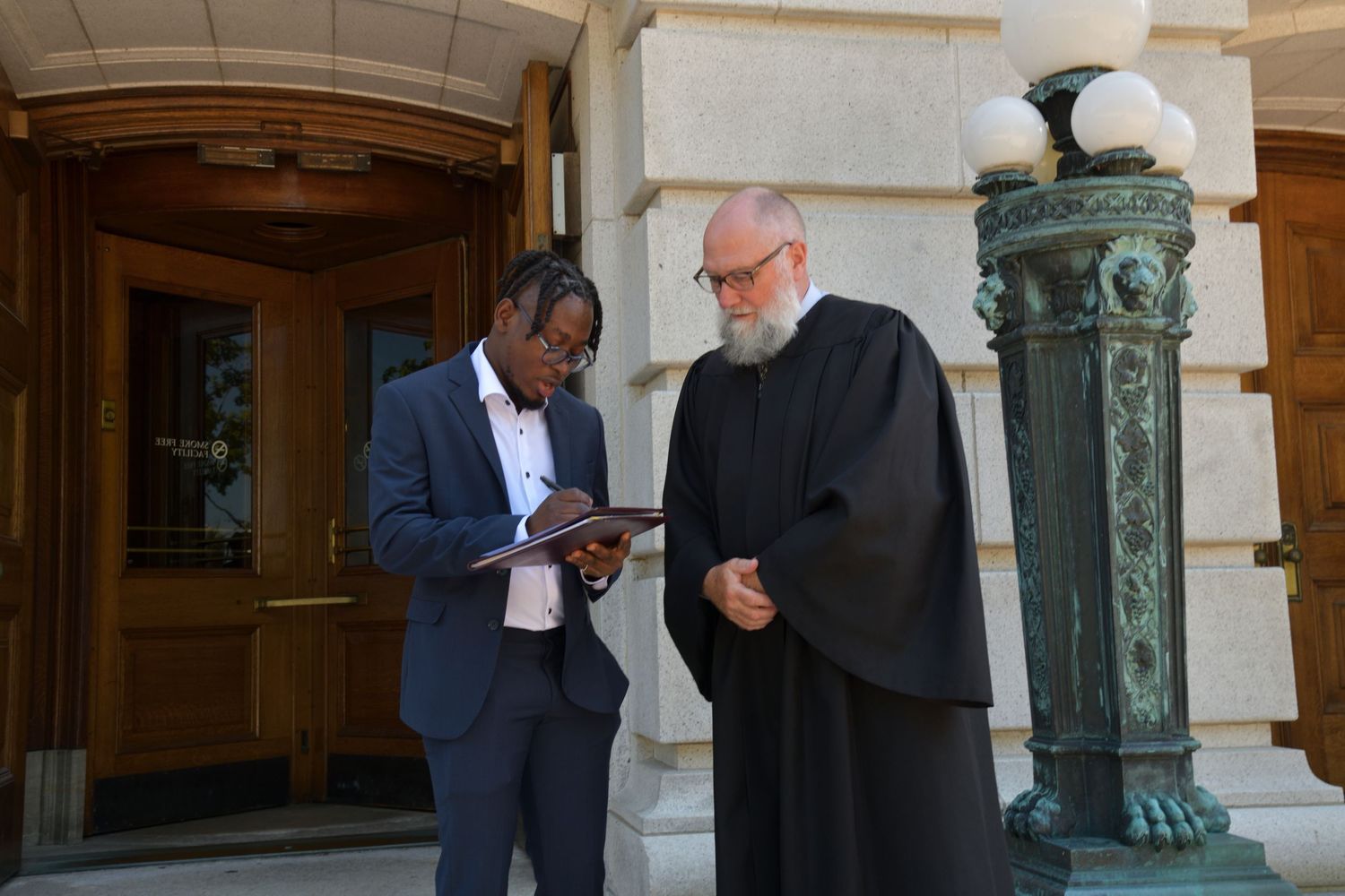 Groom in blue suit reviews wedding documents with officiant in black robes outside stone building.