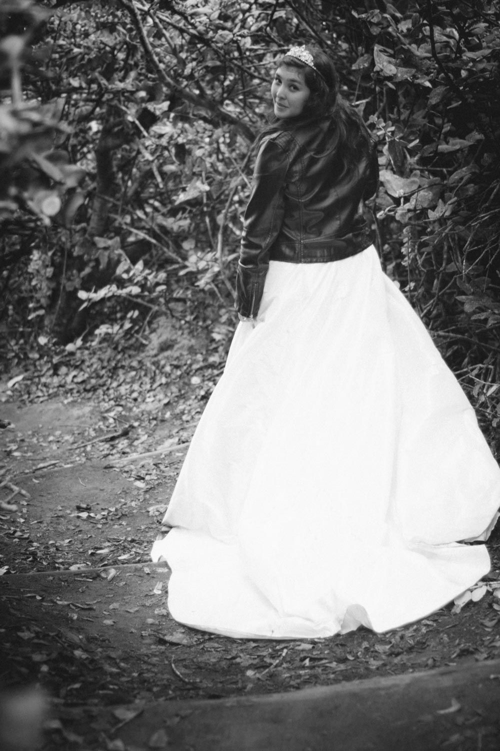 Bride in white ball gown and leather jacket poses in a forest path in black and white photo.