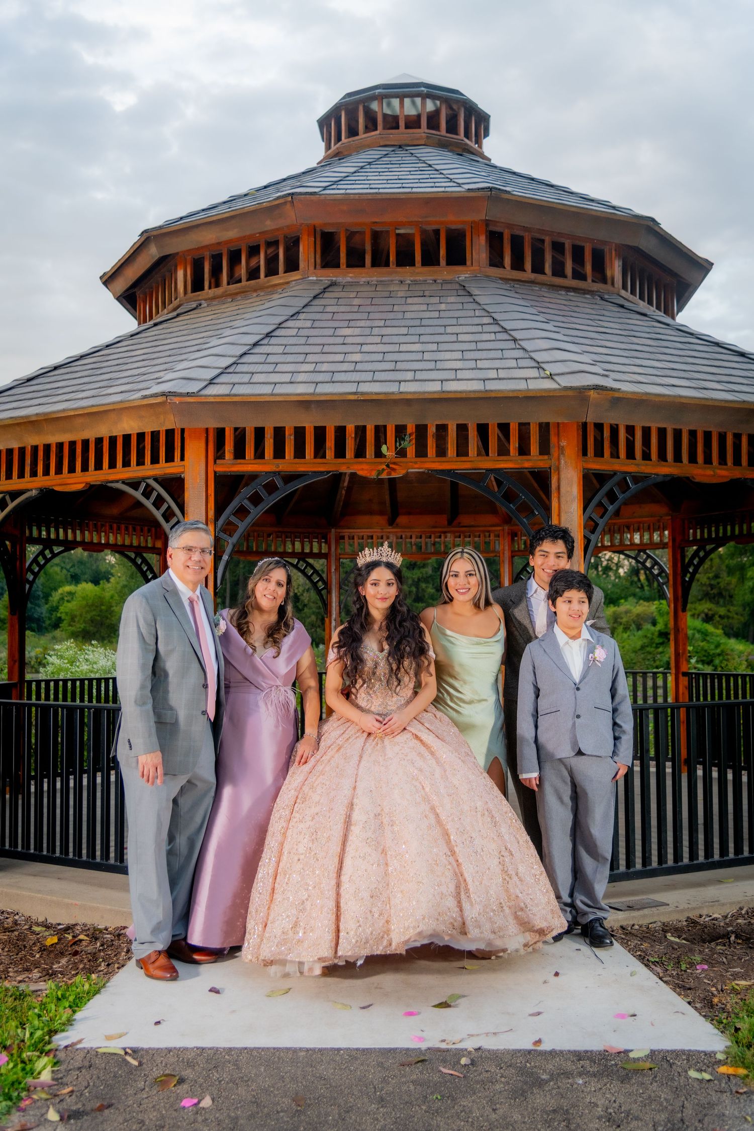 Quinceañera girl in rose gold gown poses with family of five in front of large wooden gazebo outdoors.