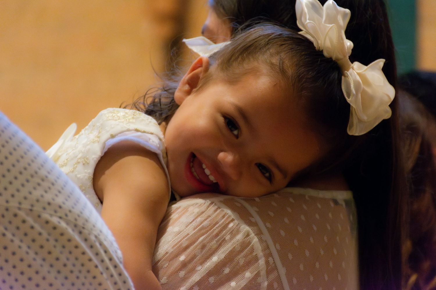 Smiling little girl with white bow resting her chin on an adult's shoulder at an indoor event.