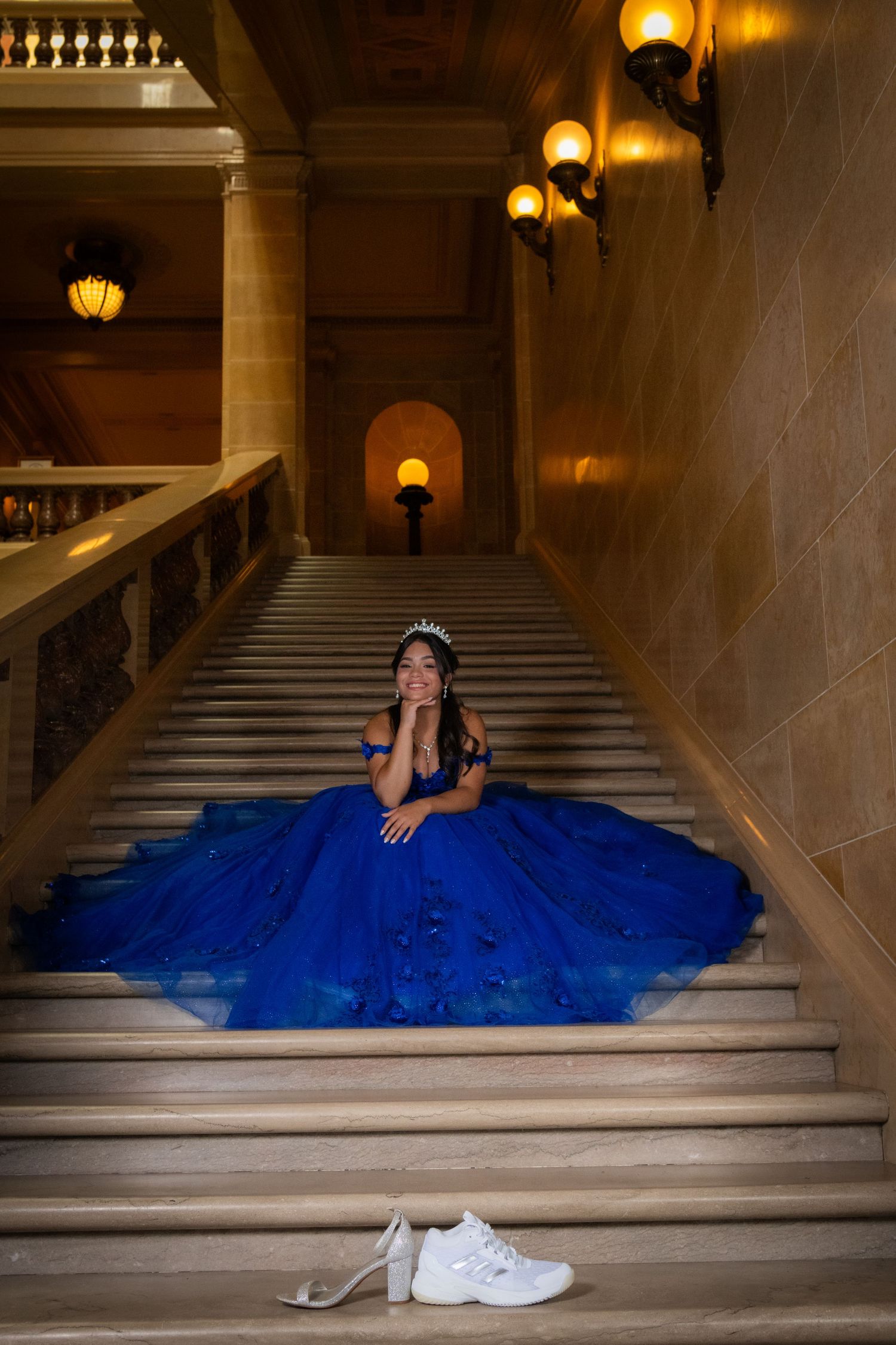 Young woman in blue quinceañera gown sitting on grand staircase with sneakers nearby.