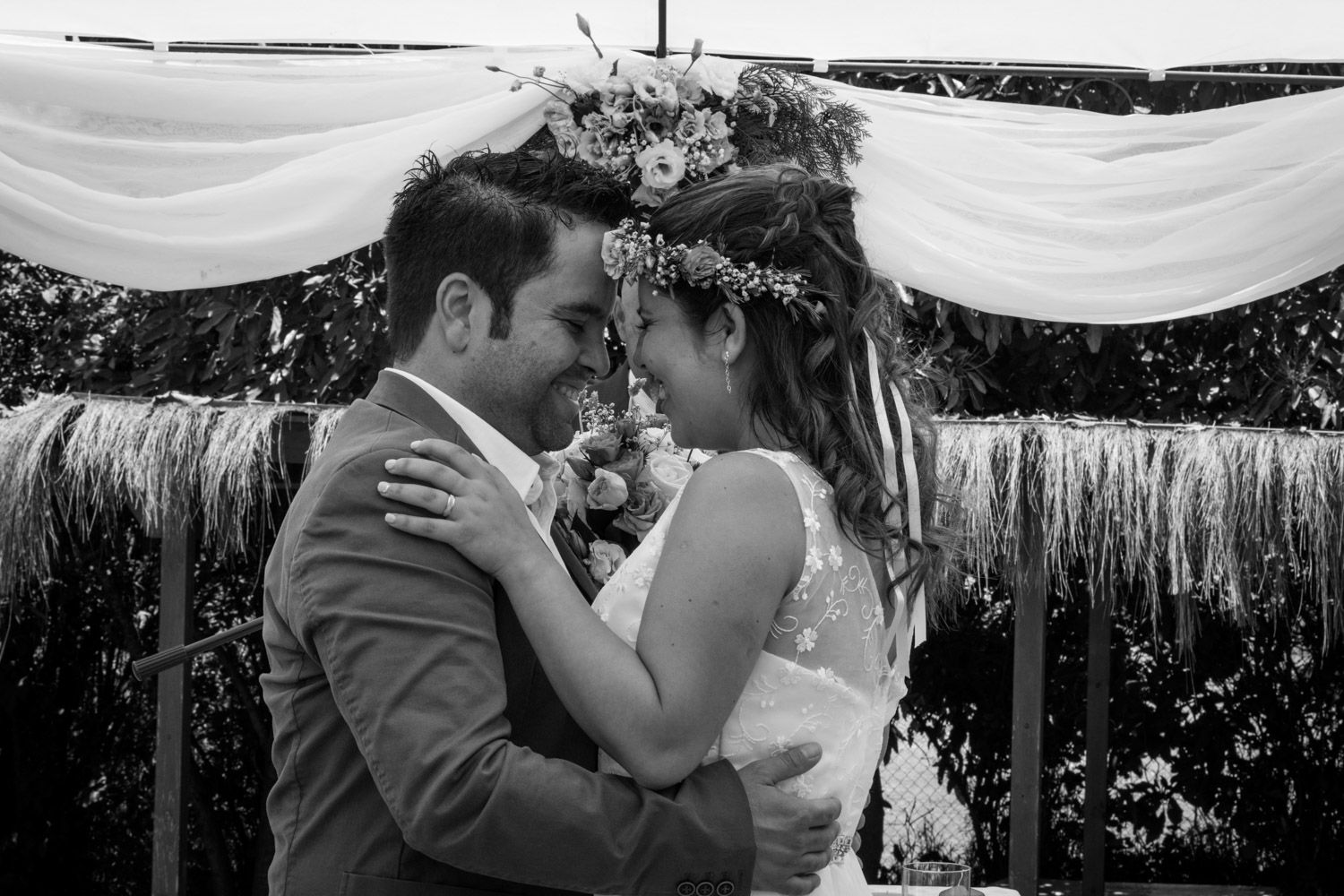 Black and white romantic wedding photo of bride and groom sharing intimate moment under floral arch.