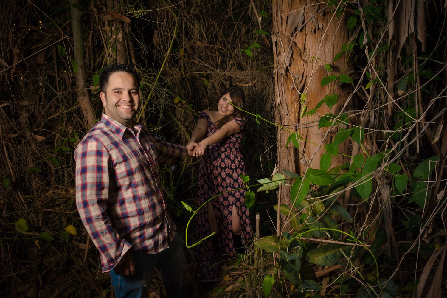 Man in plaid shirt smiling outdoors at night with woman standing behind a vine-covered tree in forest.