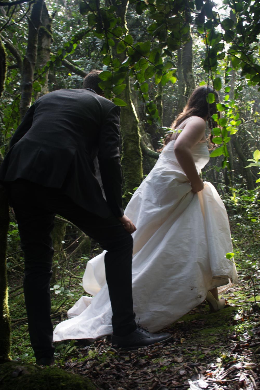 Groom assists bride with her white wedding gown train while walking through a dense forest.