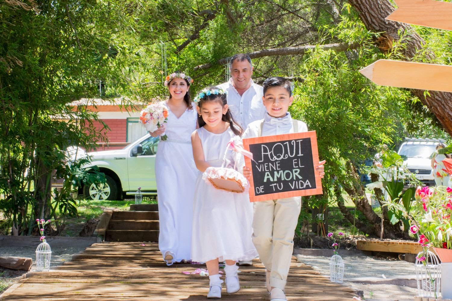 Two children in white attire holding chalkboard sign leading wedding procession through outdoor garden venue.
