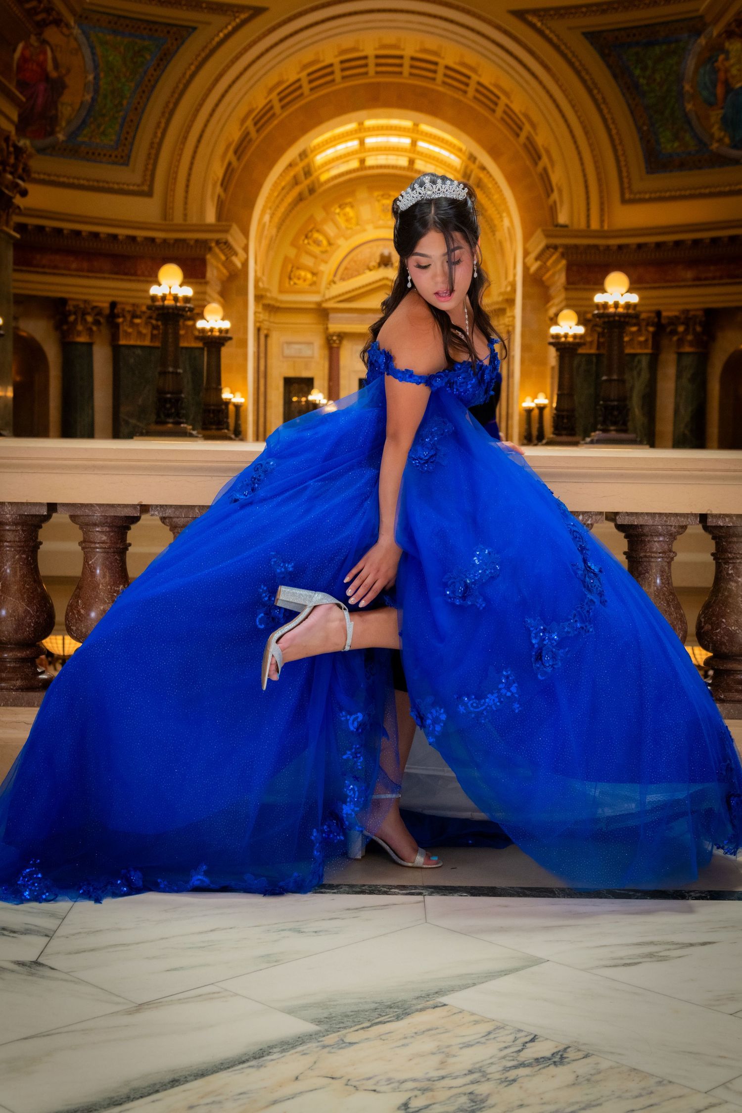 Woman in royal blue quinceañera ball gown adjusting heels in grand ornate hall.