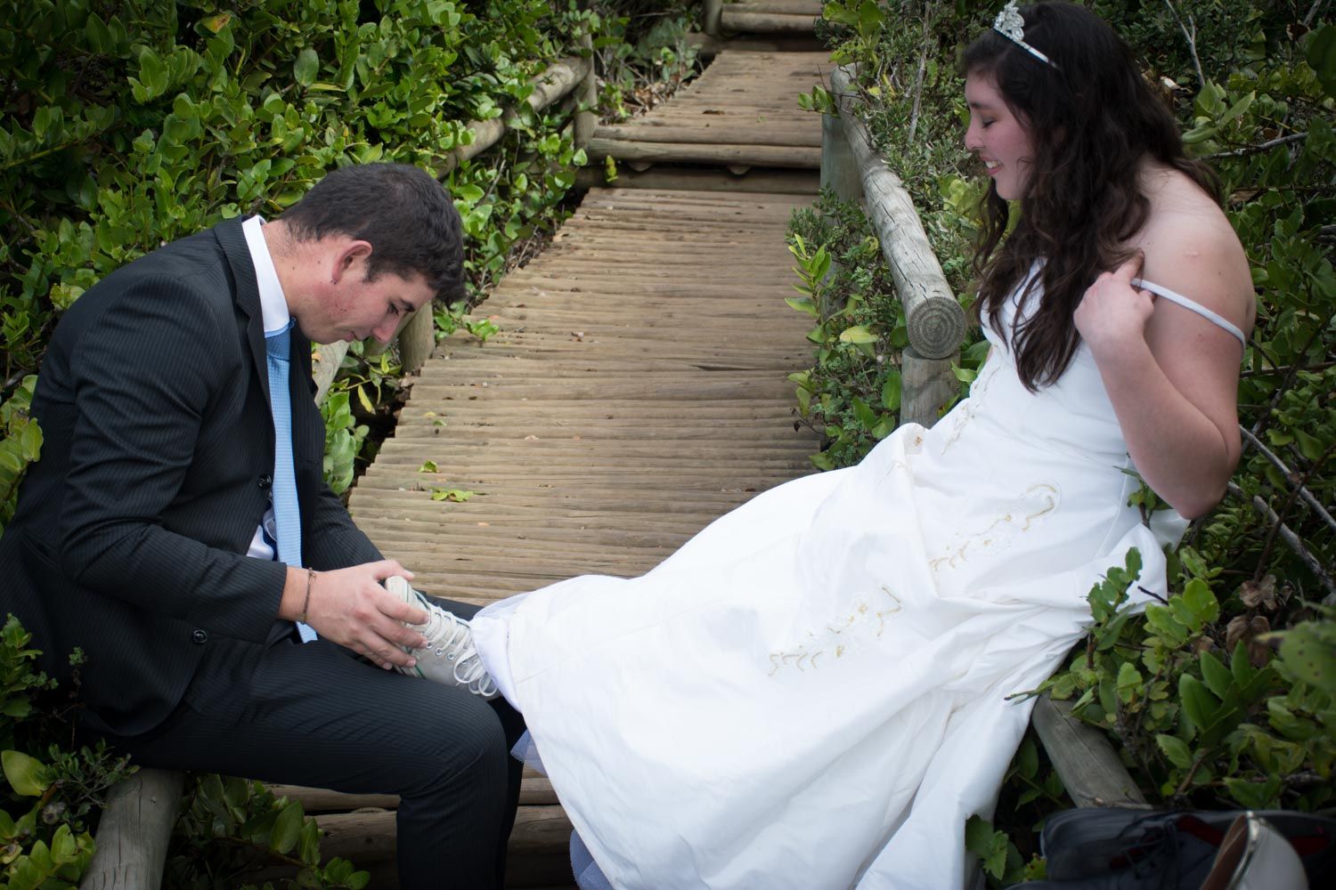Groom tenderly adjusting brides shoe on rustic wooden boardwalk surrounded by greenery.