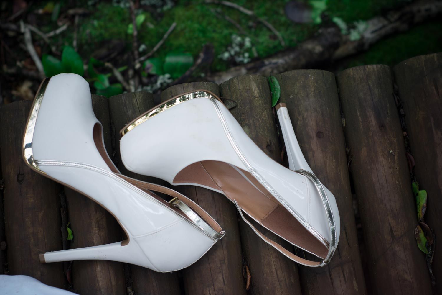 White patent leather high heel shoes with gold trim resting on rustic wooden logs outdoors.