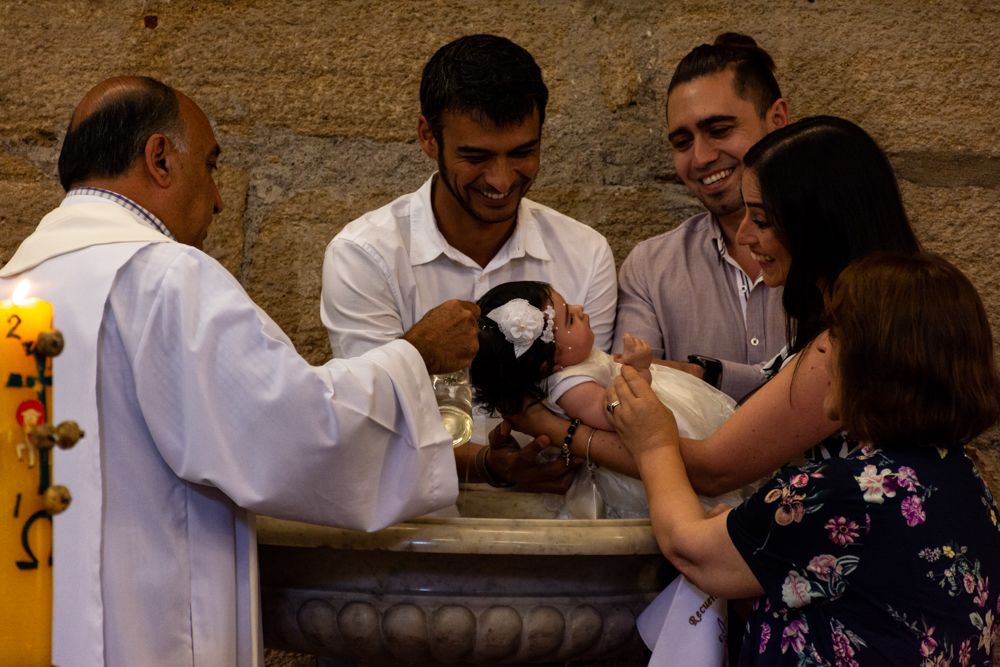 Priest baptizes infant at stone font surrounded by smiling family members in a church.
