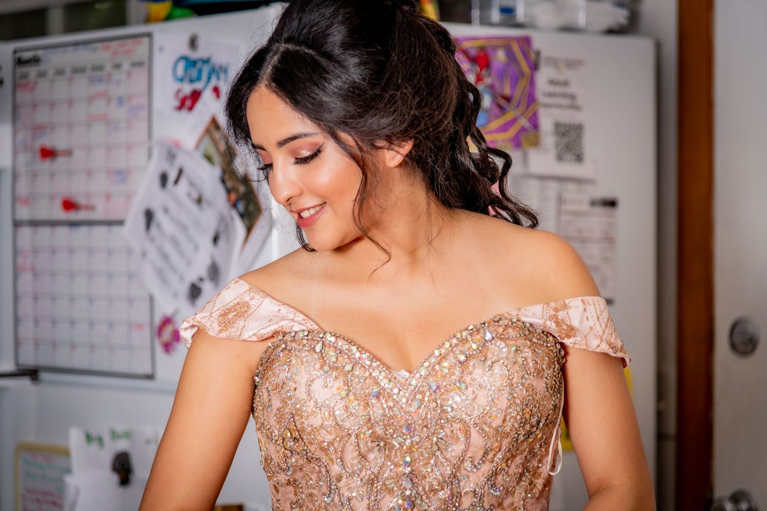 Young woman in elegant off-shoulder gold embroidered formal gown smiling downward indoors.