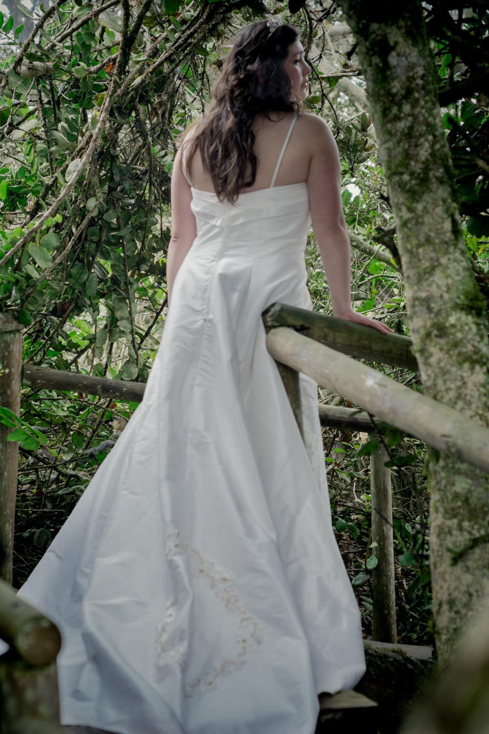 Bride in elegant white wedding gown standing on wooden staircase surrounded by lush greenery.