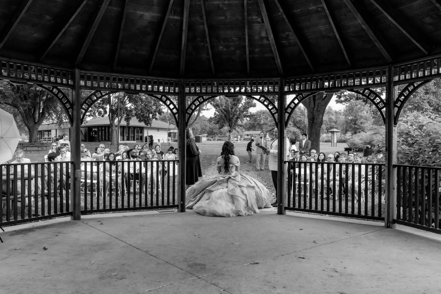 Black and white photo of quinceañera girl in voluminous ballgown inside a classic wooden gazebo structure.
