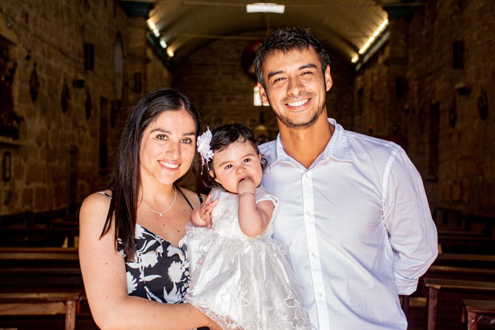 Happy parents holding their baby dressed in a white christening gown inside a historic stone church.
