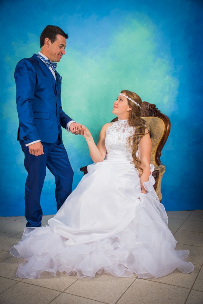 Groom in blue suit holds bride's hand as she sits in ornate chair against blue painted wall.