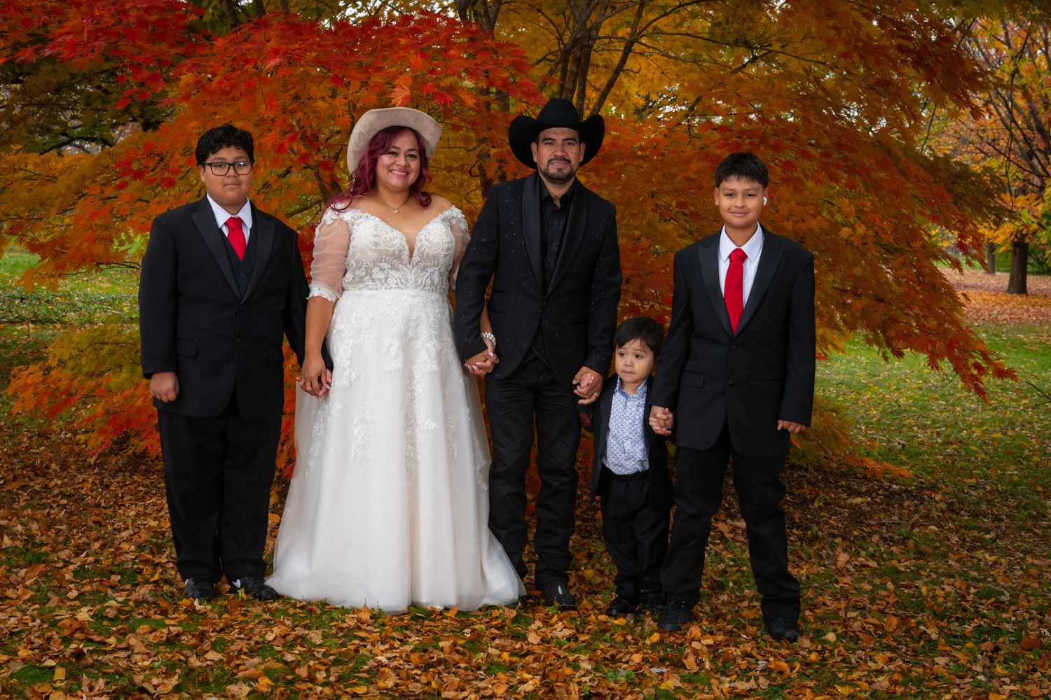 Bride and groom with three sons posing before vibrant red maple tree in fall park.