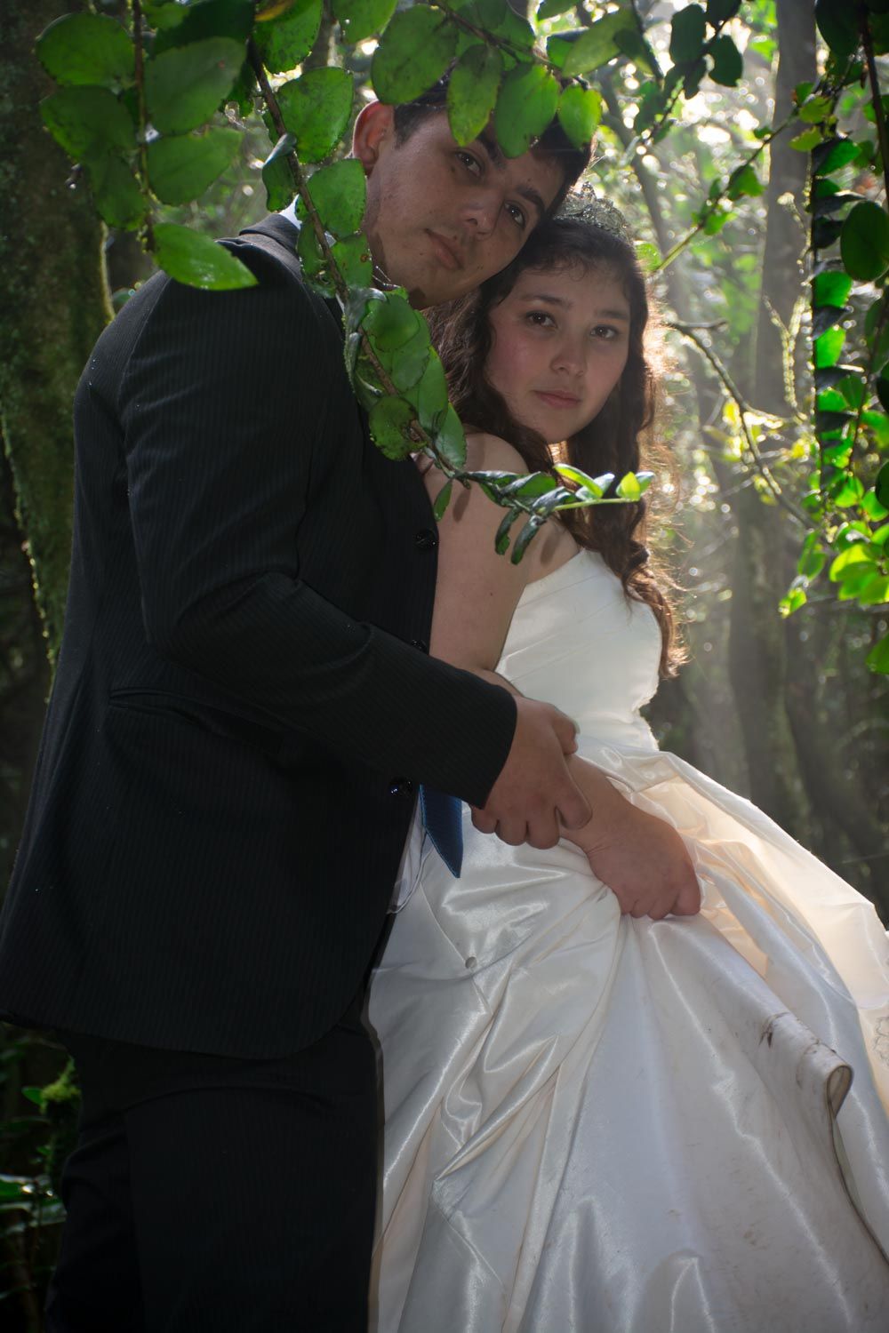 Bride and groom pose together under green leaves in a sunlit forest setting.