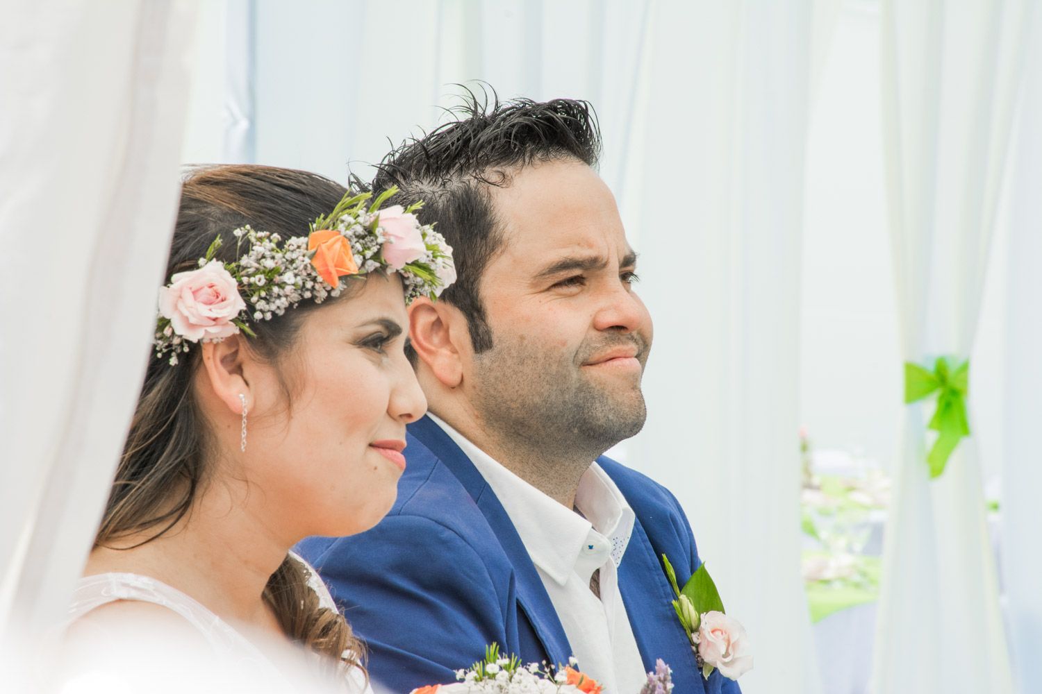 Close-up of bride wearing pink floral crown and groom in blue suit smiling during outdoor wedding ceremony.