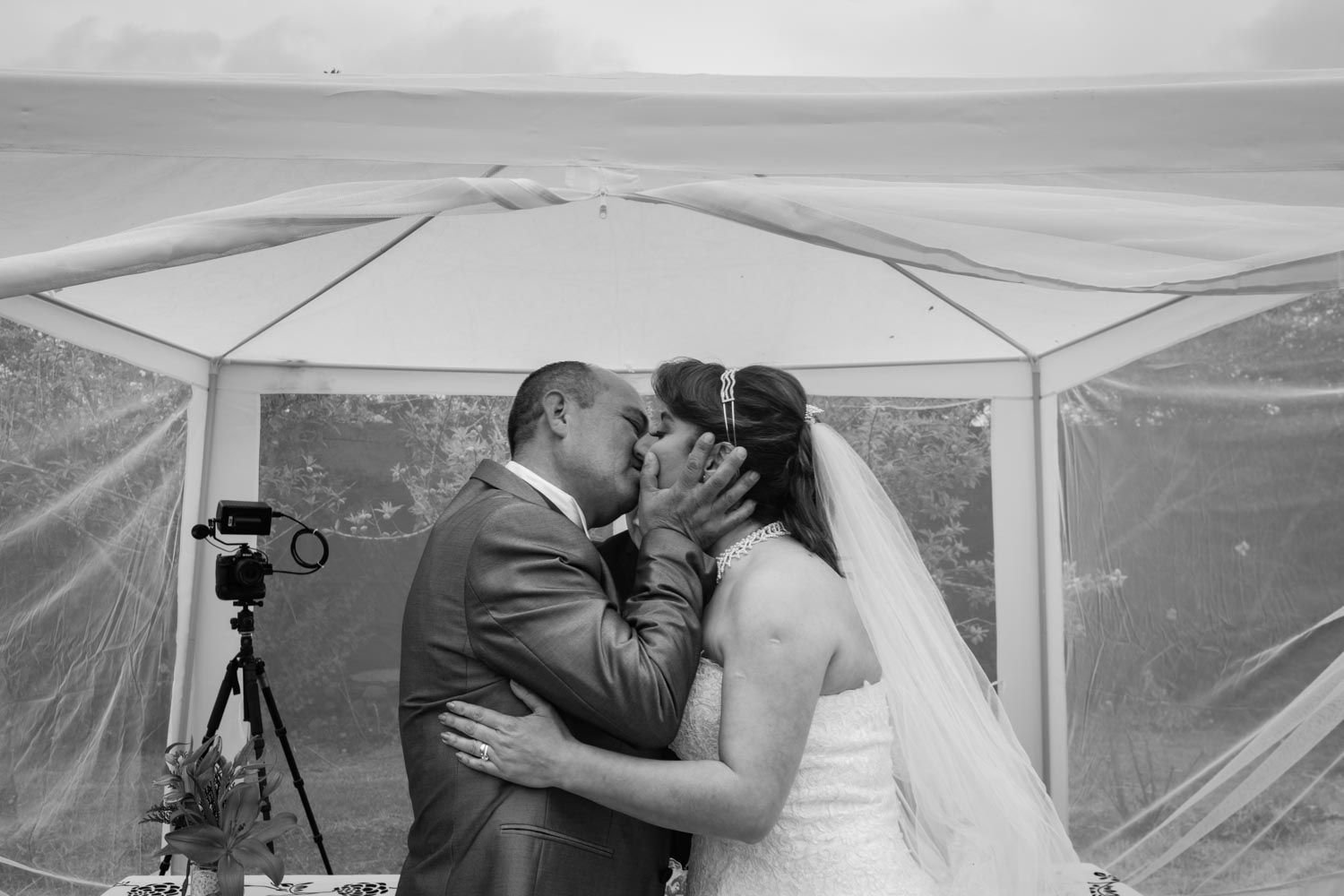 Black and white photo of bride and groom sharing first kiss under wedding canopy by the ocean.