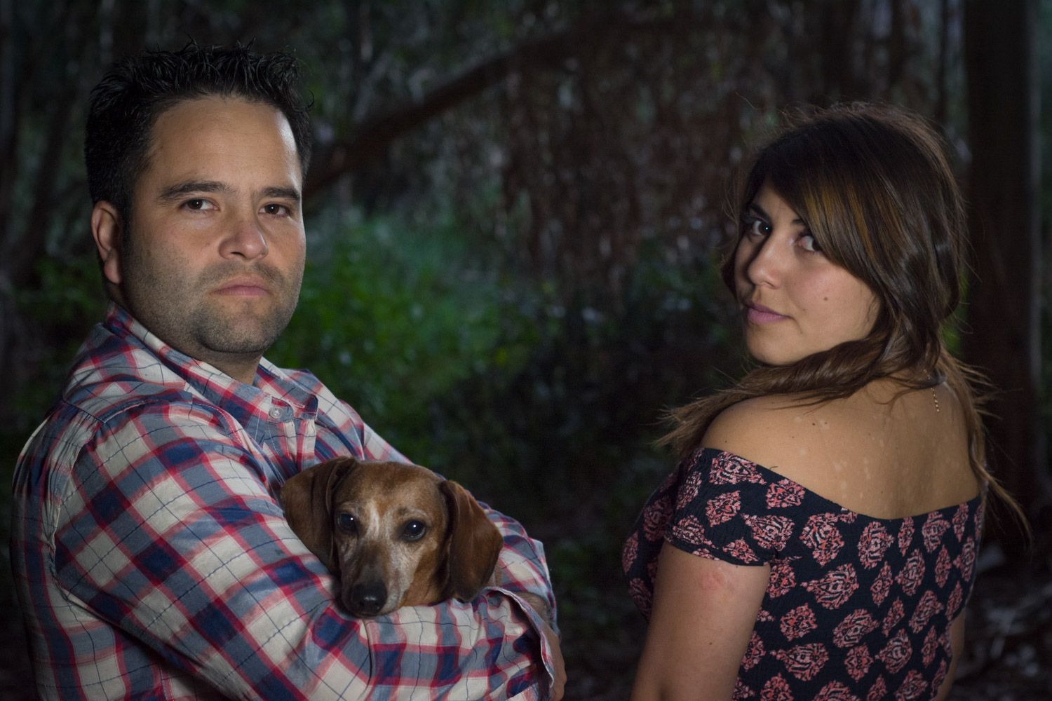 Man holding a small dachshund dog with arms crossed while woman stands nearby in a dark forest setting.