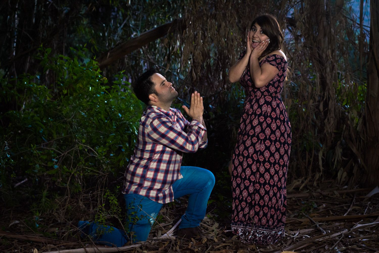 Man on one knee proposing to surprised woman covering her face with hands in a romantic forest setting.