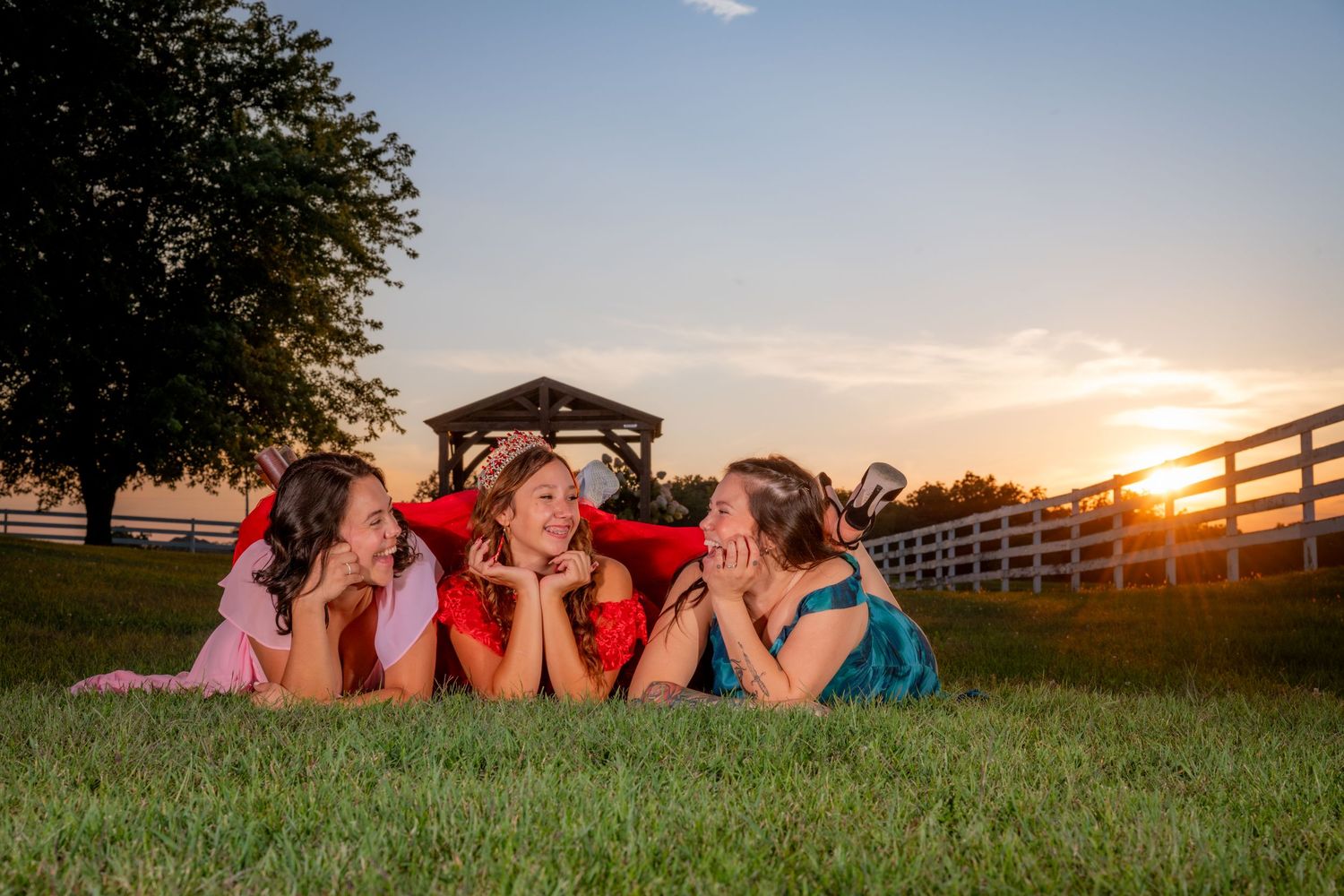 Three girls in colorful dresses lie on green grass laughing together at a golden sunset.
