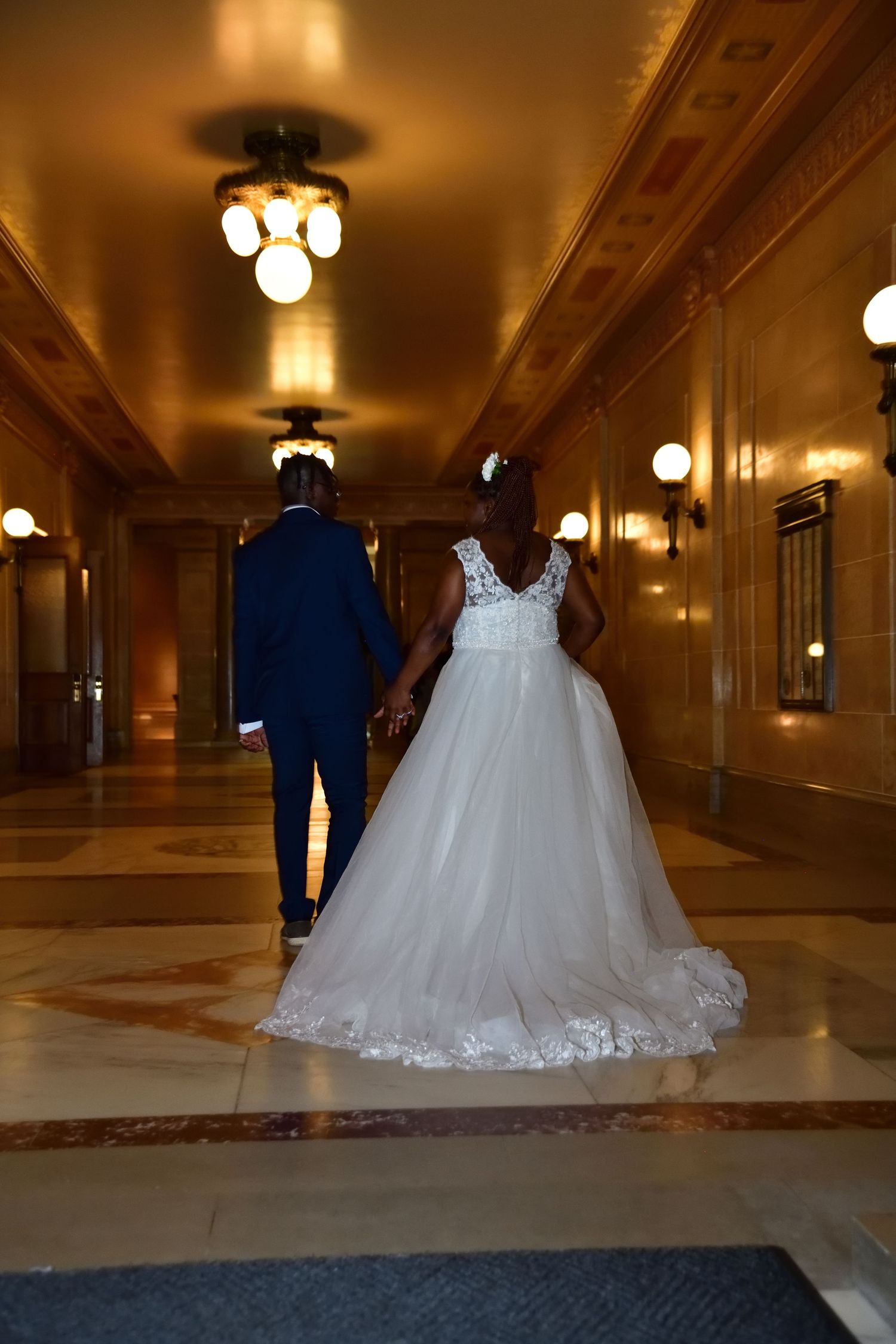 Newlyweds walk hand in hand down ornate golden hallway after wedding ceremony.