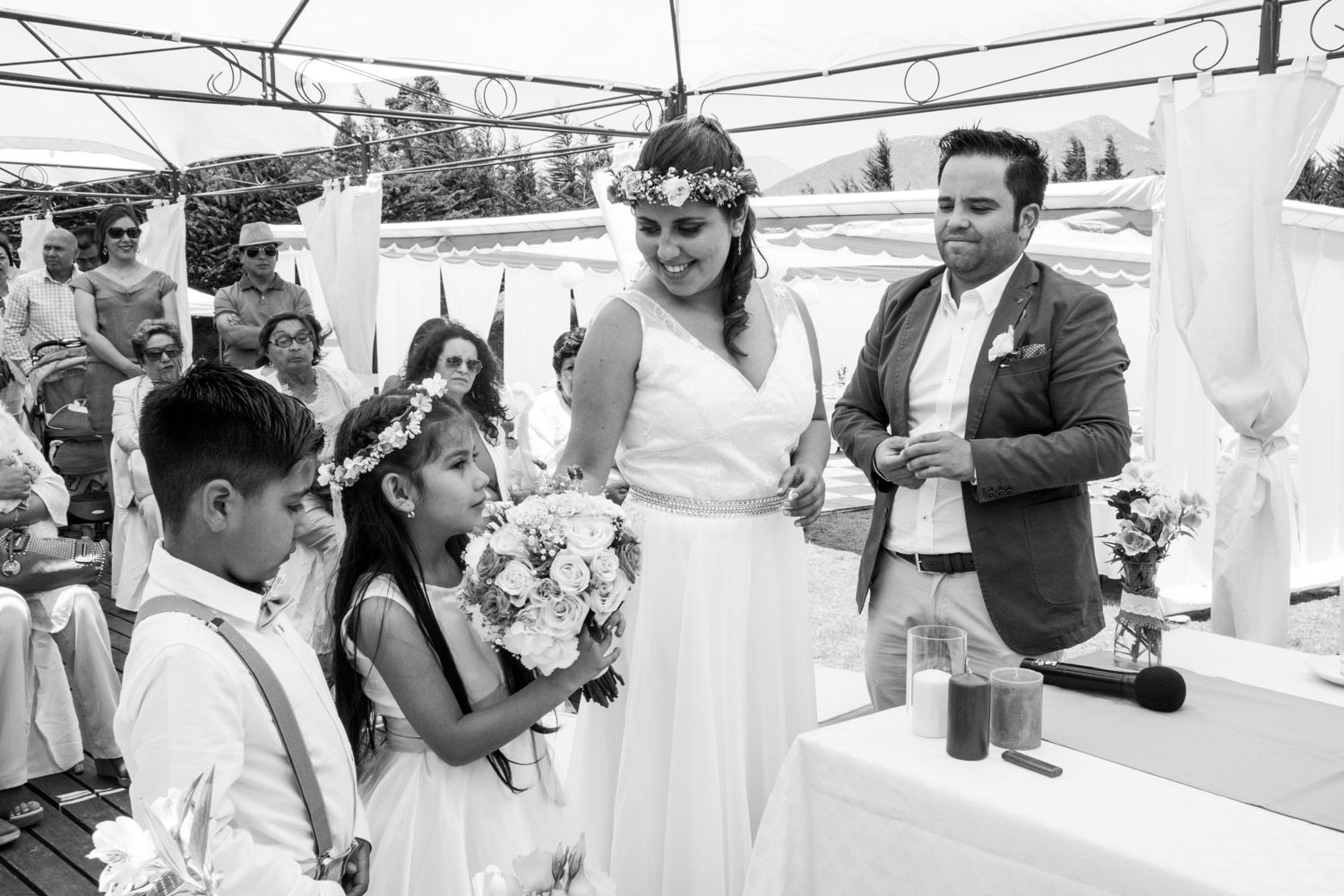 Bride in floral crown laughing with flower girl and ring bearer during black and white outdoor wedding ceremony.