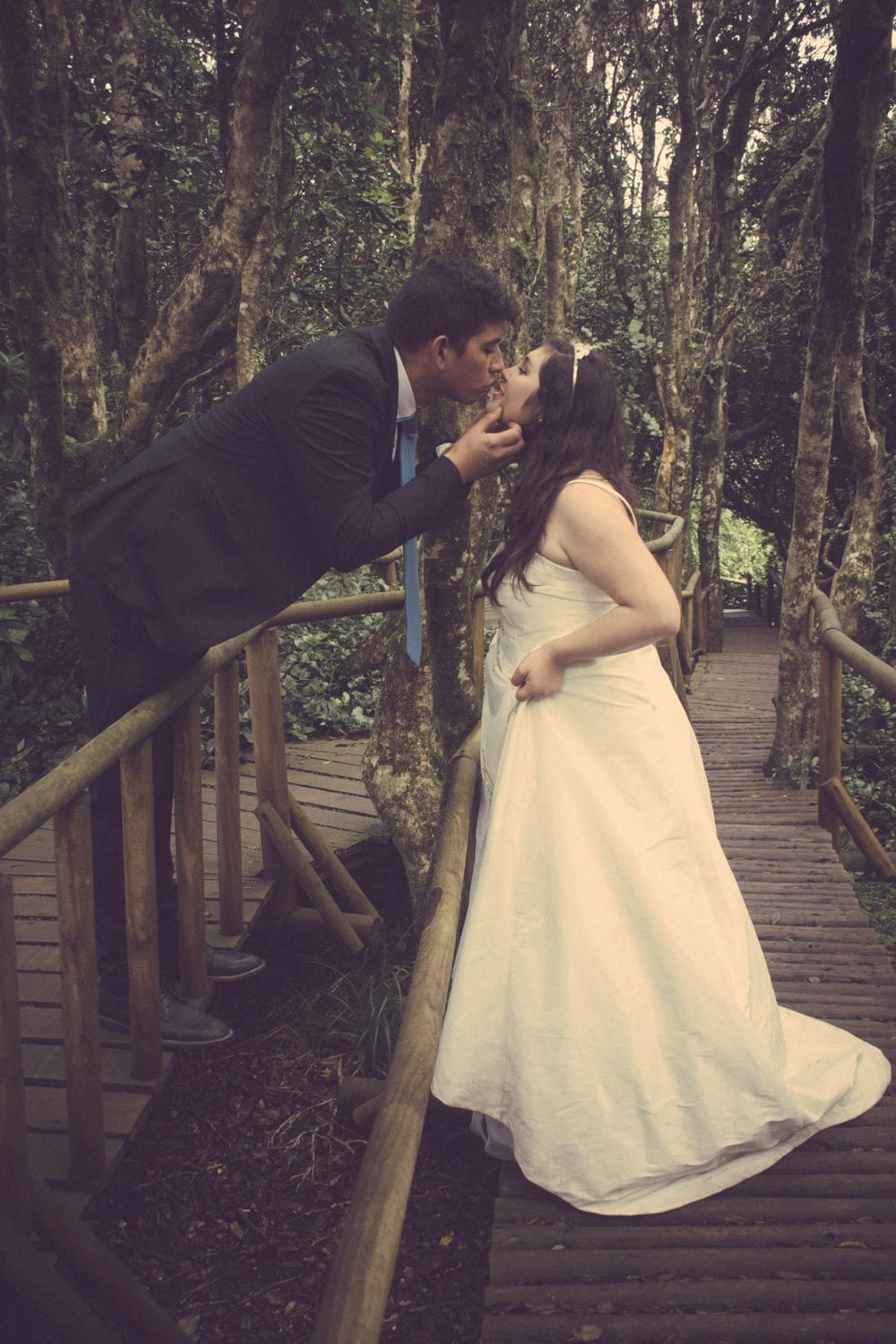 Bride and groom sharing a romantic kiss on a rustic wooden bridge surrounded by lush forest trees.