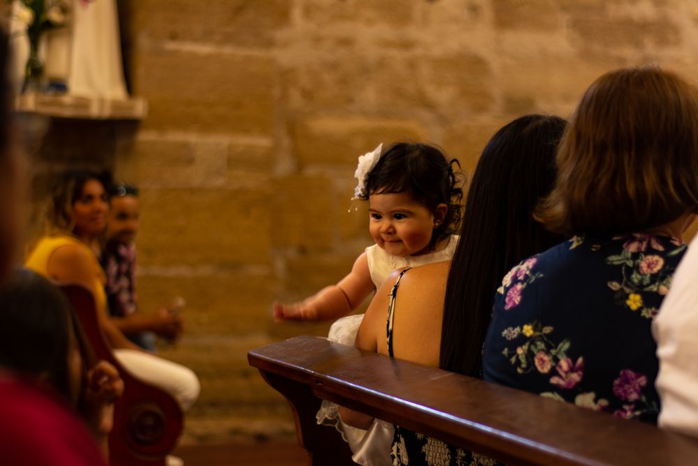 A smiling toddler in white dress sits on church pew during a religious ceremony with family members nearby.