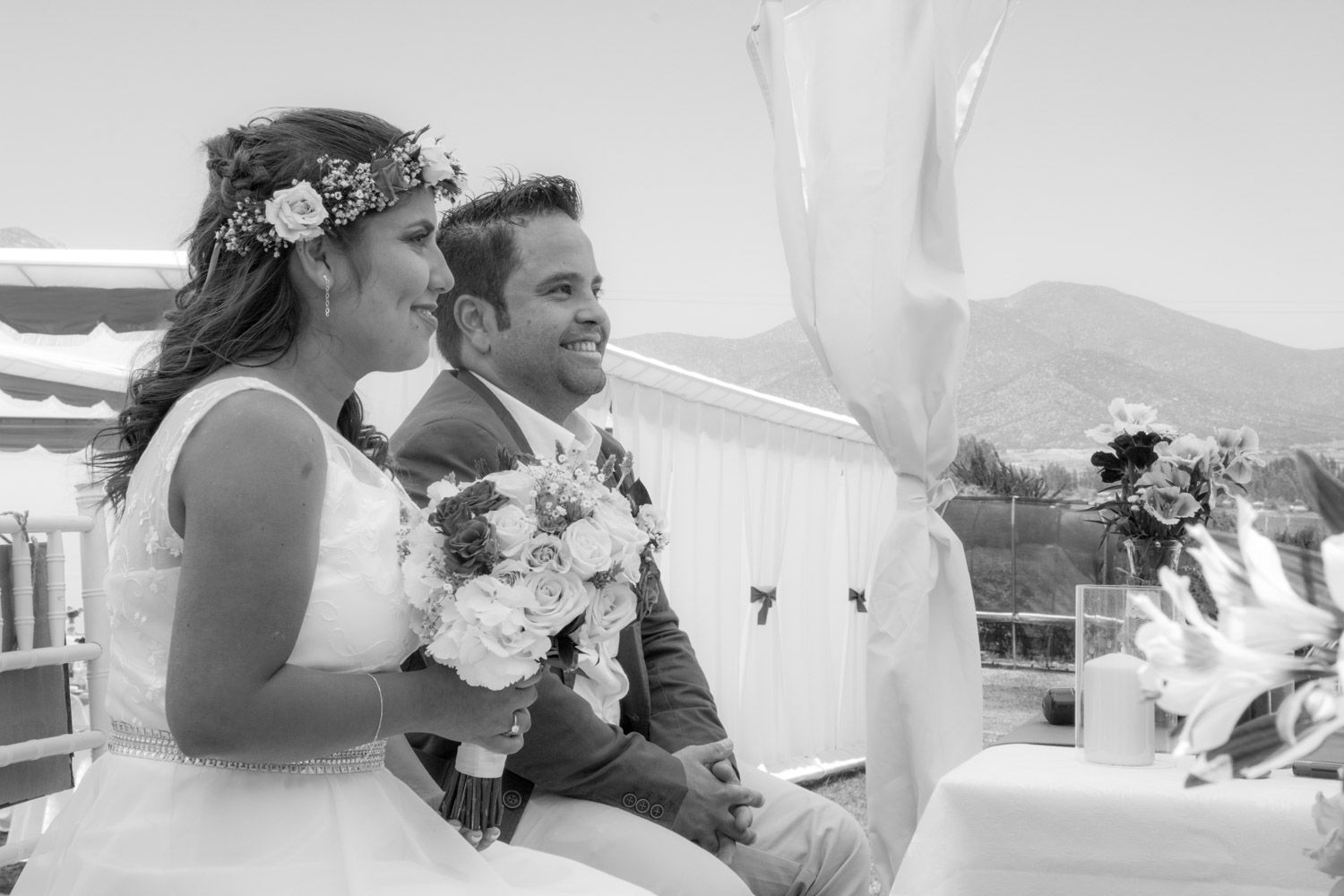 Black and white photo of bride with flower crown and groom seated together during outdoor wedding ceremony.