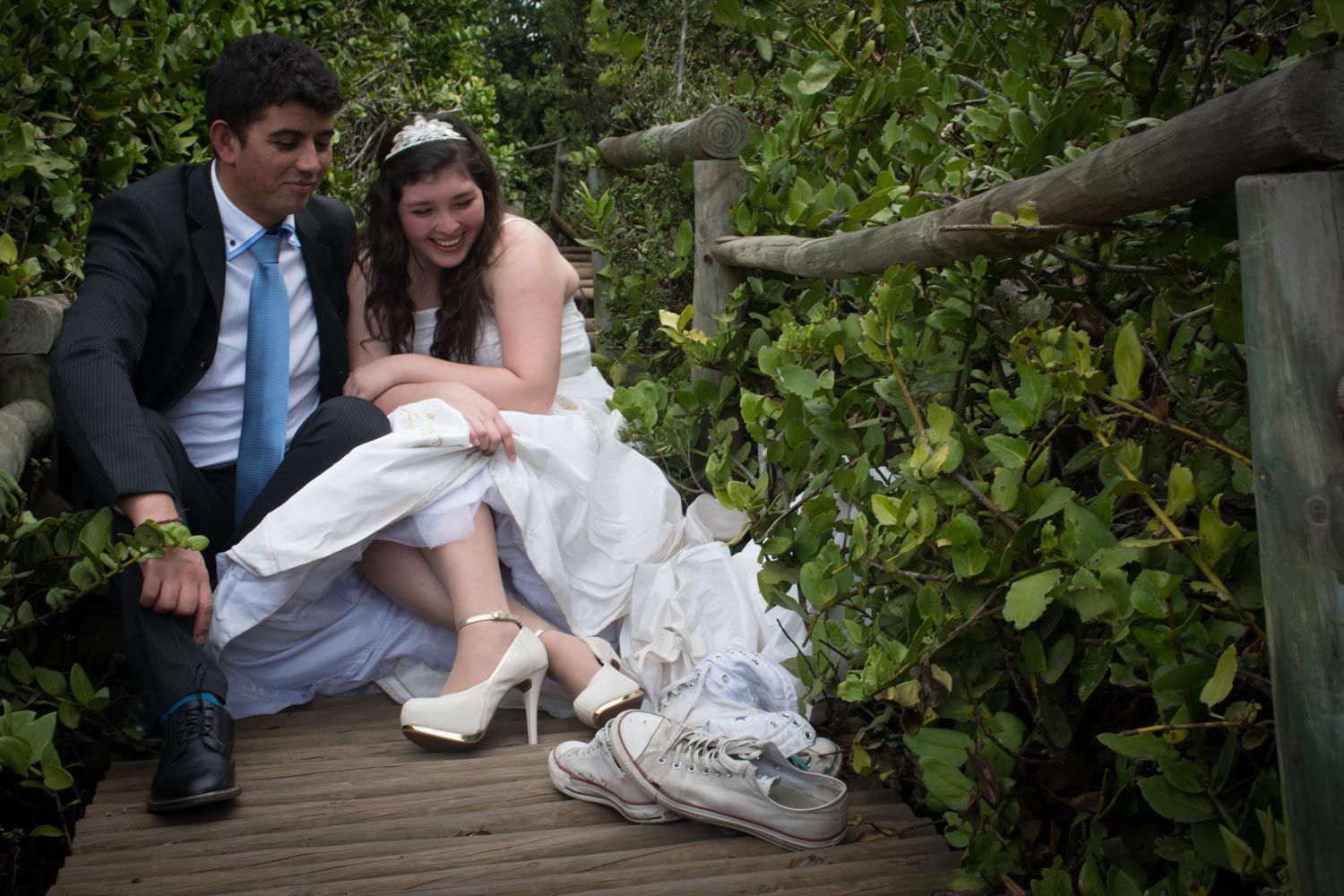 Groom helps bride change shoes on a rustic wooden bridge surrounded by green bushes.