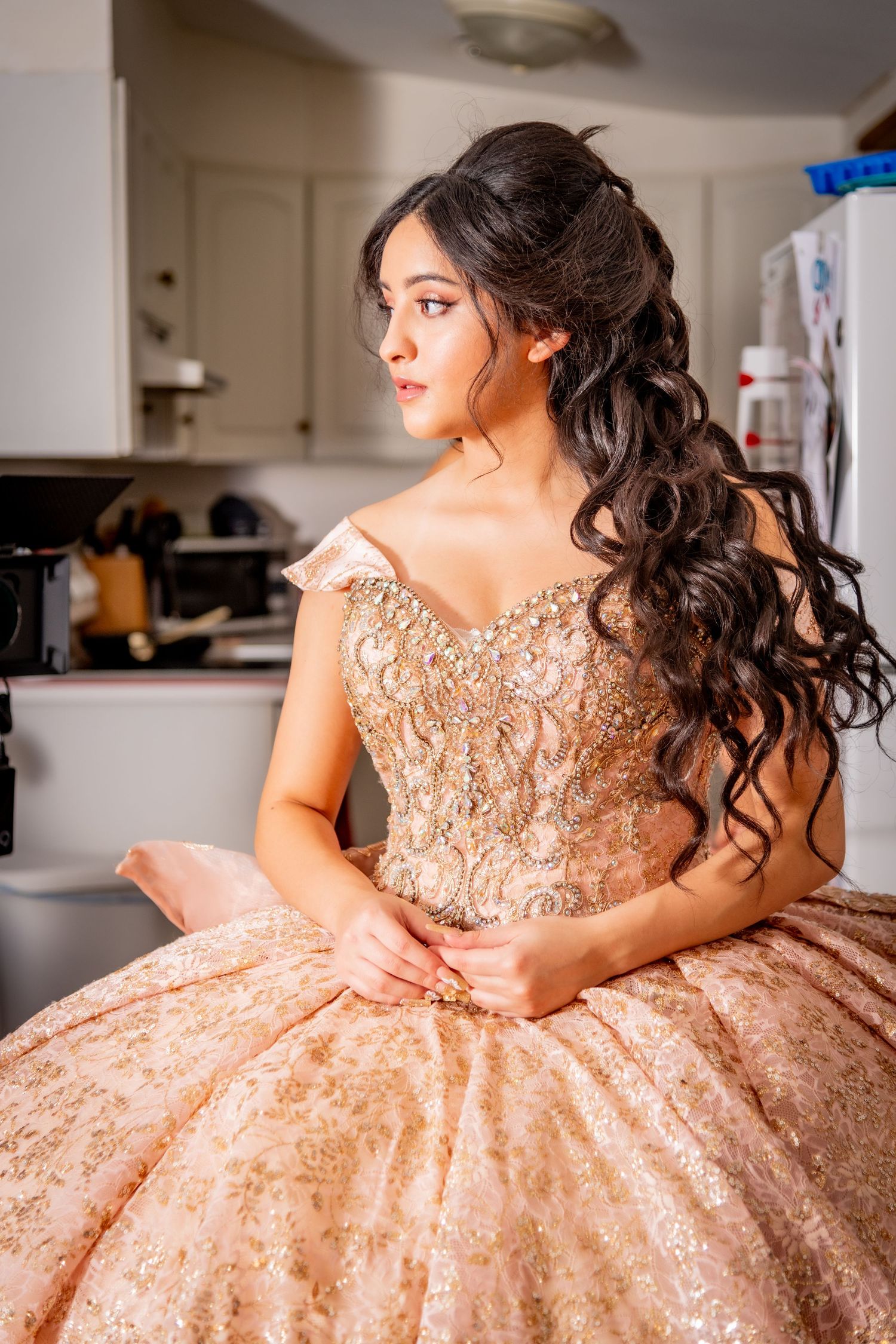 Young woman in rose gold quinceañera ballgown with intricate beading sits gracefully in a kitchen setting.