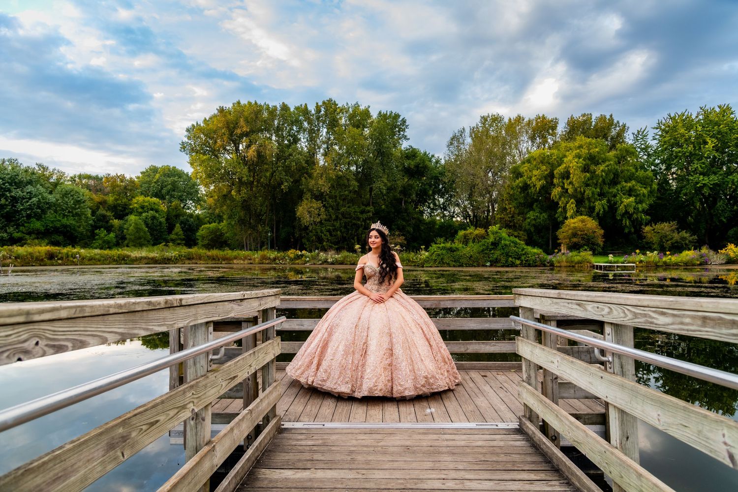 Young woman in a stunning pink quinceañera ball gown posing on a wooden bridge in a park.