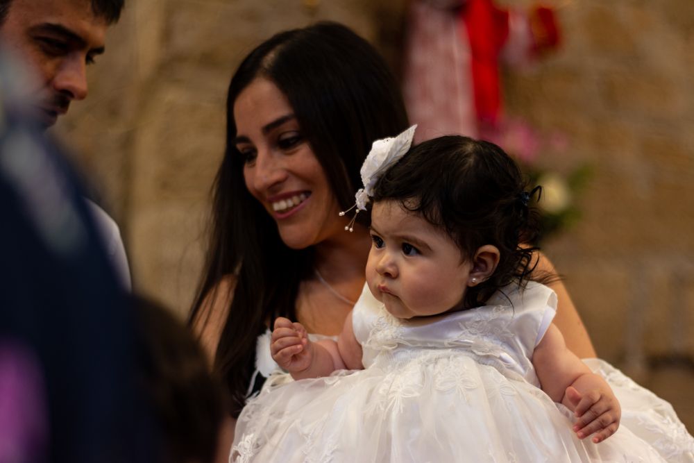 Smiling woman in white dress holding a baby girl in a white gown at a christening ceremony.
