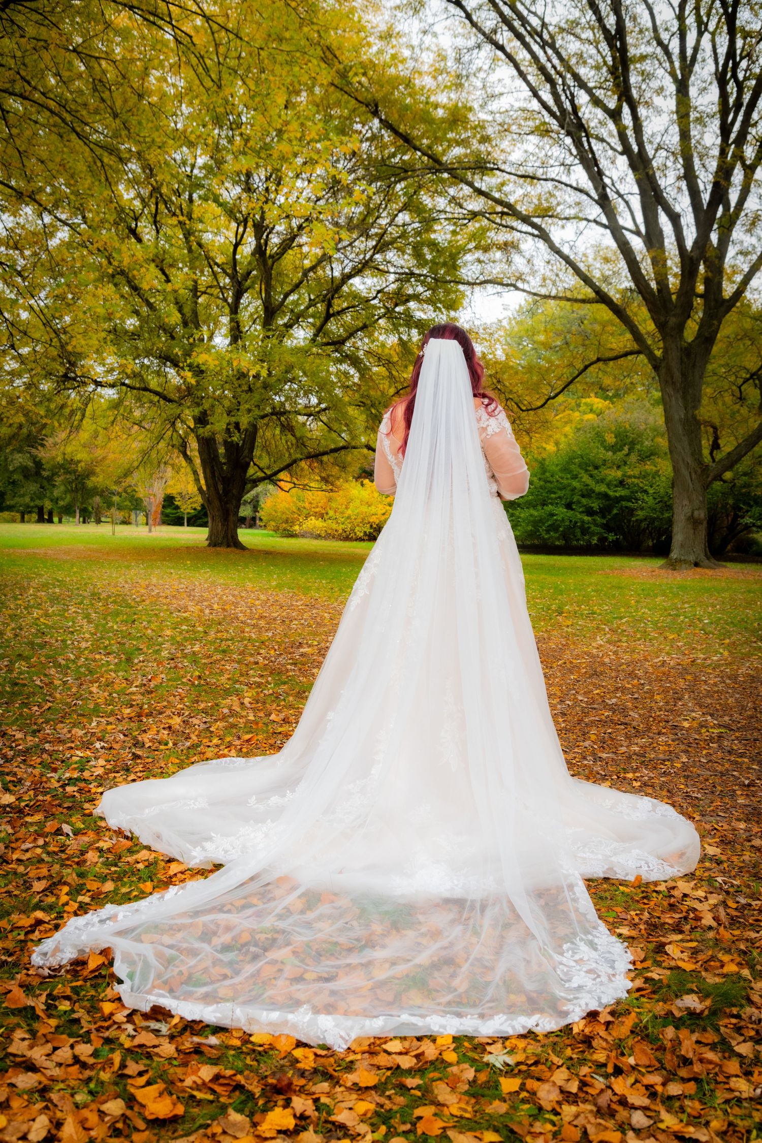 Bride with red hair and long lace veil stands in autumn park surrounded by colorful fallen leaves and trees.
