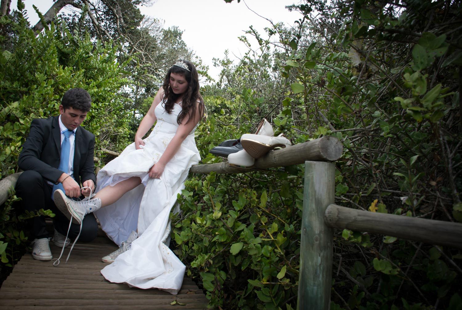 Bride and groom changing shoes on wooden boardwalk path surrounded by lush green foliage.