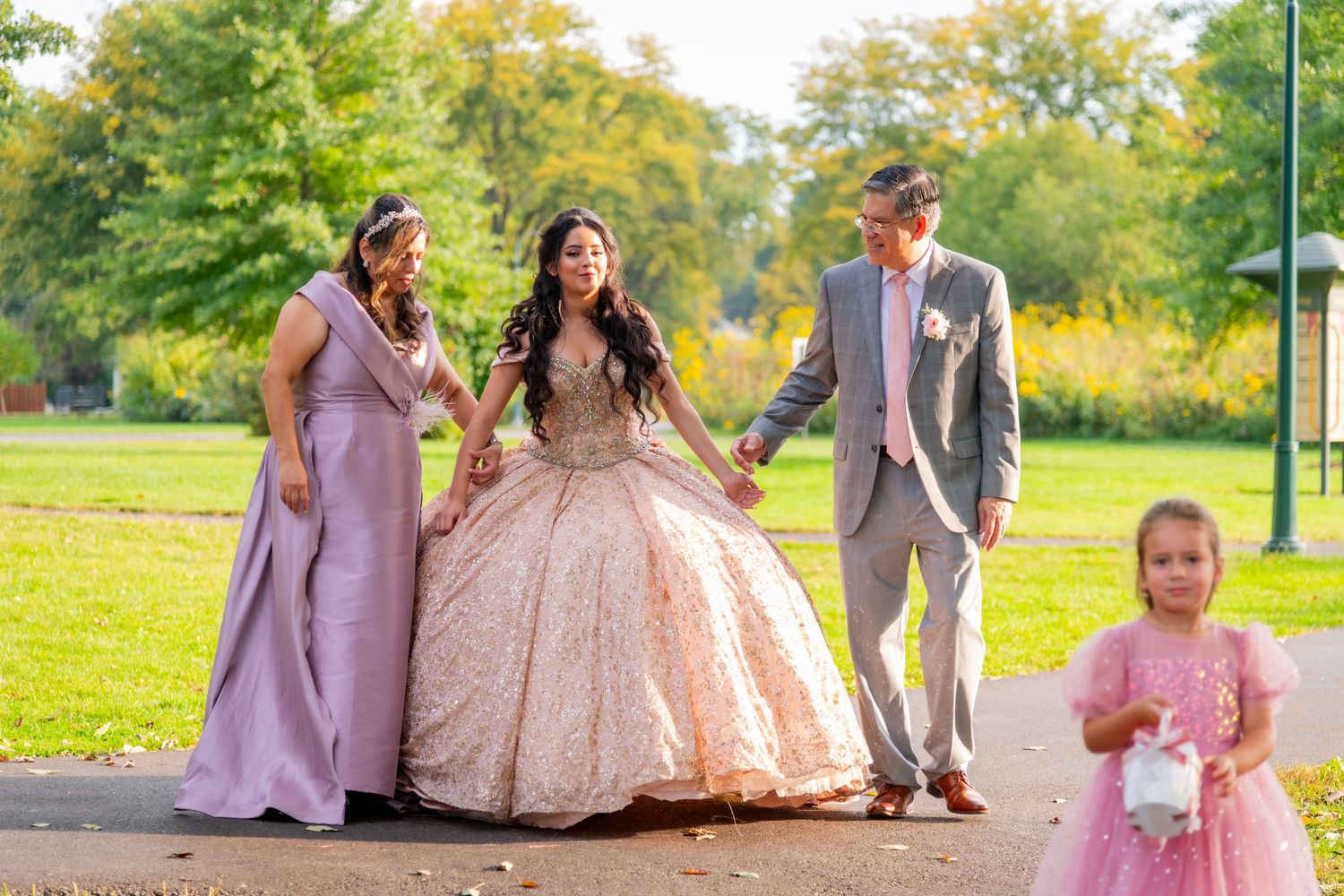 Quinceañera girl in rose gold ballgown walks with family escorts and flower girl in outdoor park setting.
