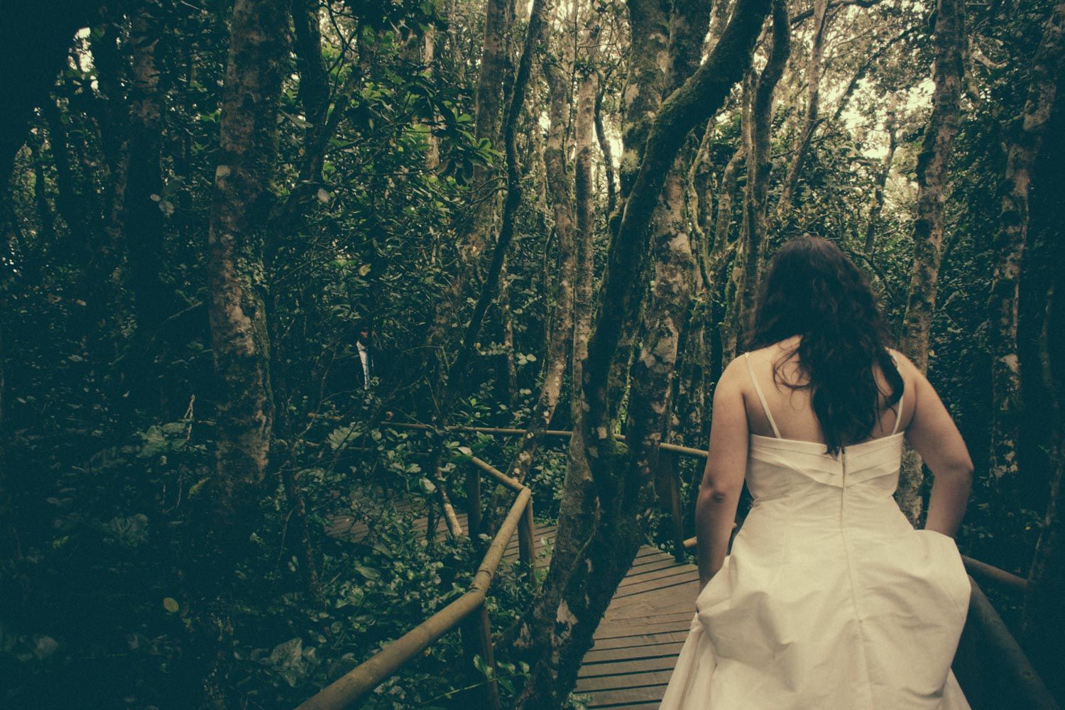 Bride in white wedding dress walking on wooden boardwalk path through lush green forest.