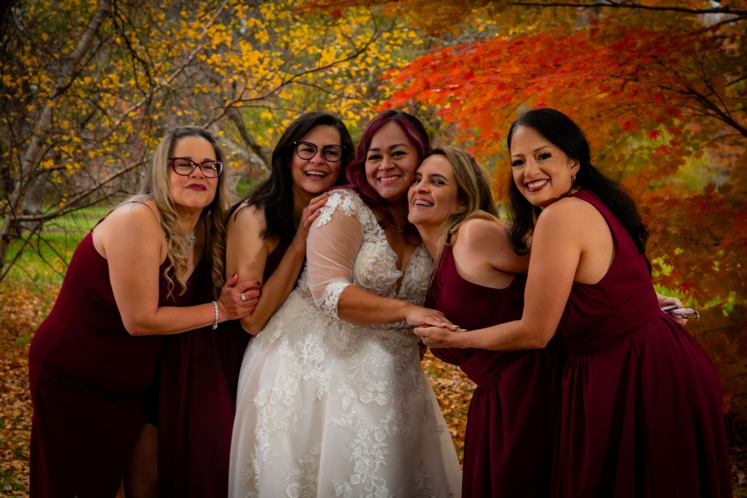 Bride in lace gown surrounded by smiling bridesmaids in burgundy dresses amid fall foliage.