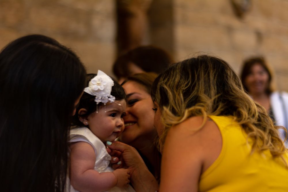 Woman in yellow dress kisses baby wearing white bow at christening celebration in church.