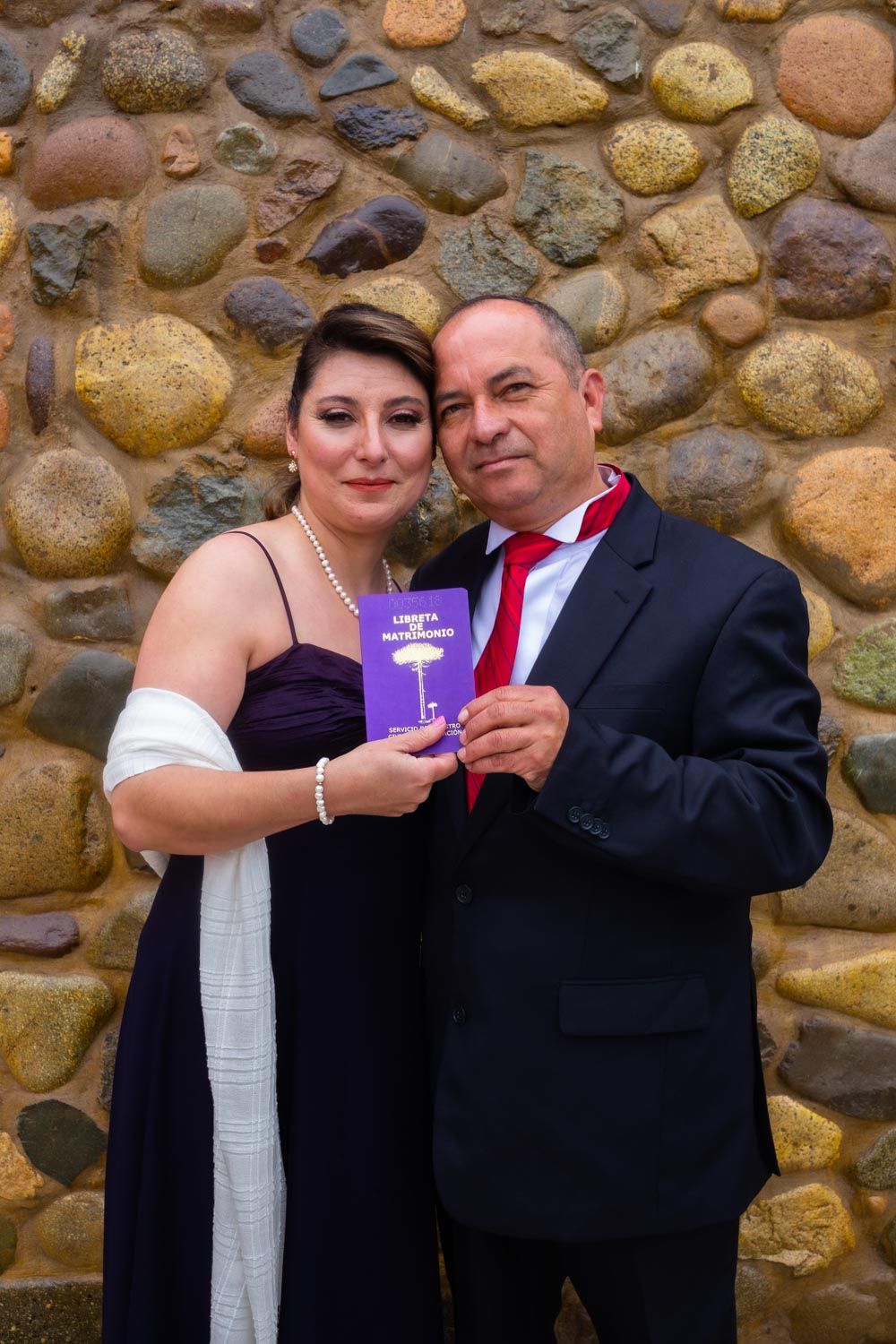 Couple in formal attire holding a purple book against a stone wall background.