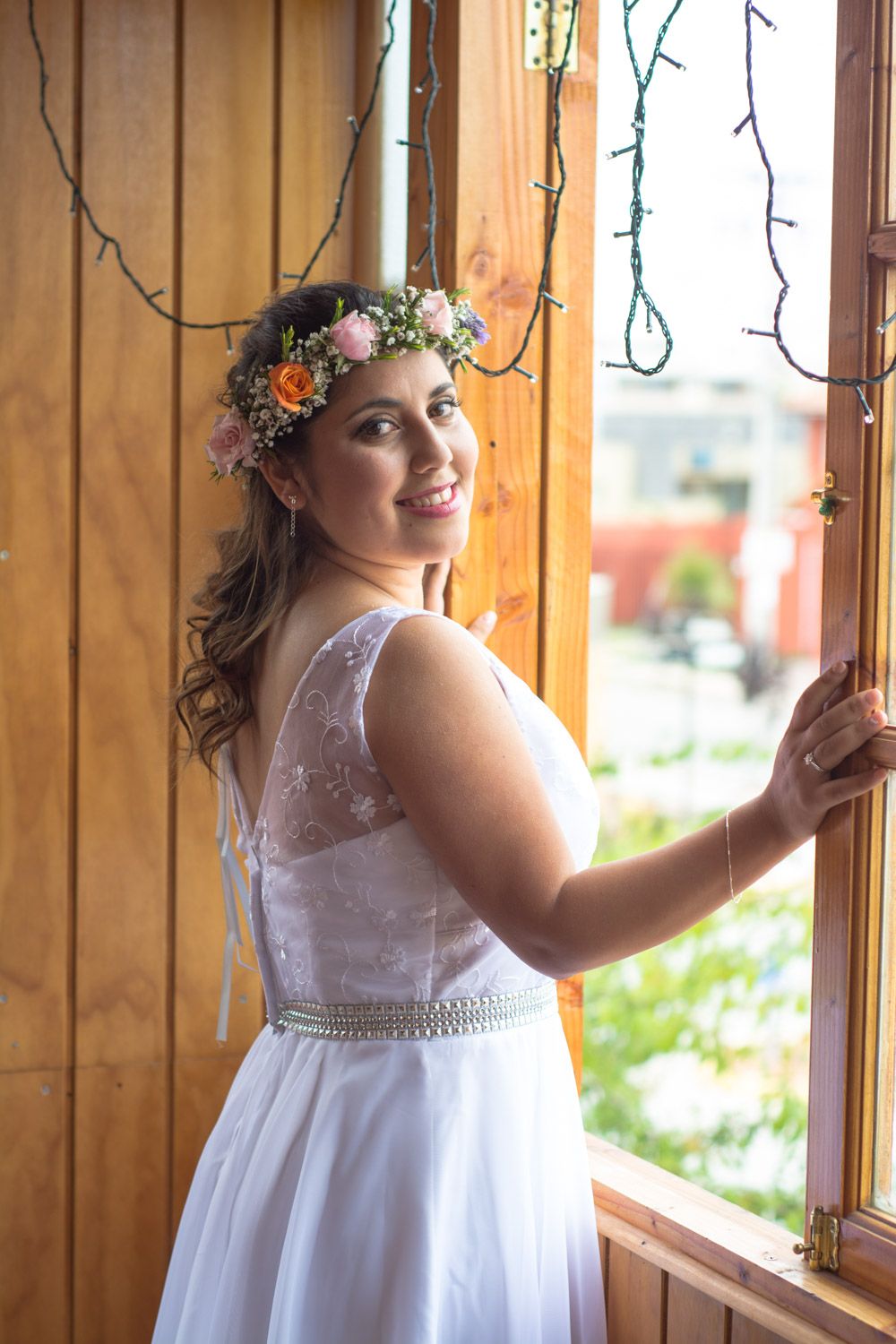 Smiling bride wearing floral crown and white lace wedding dress standing by wooden window with string lights.