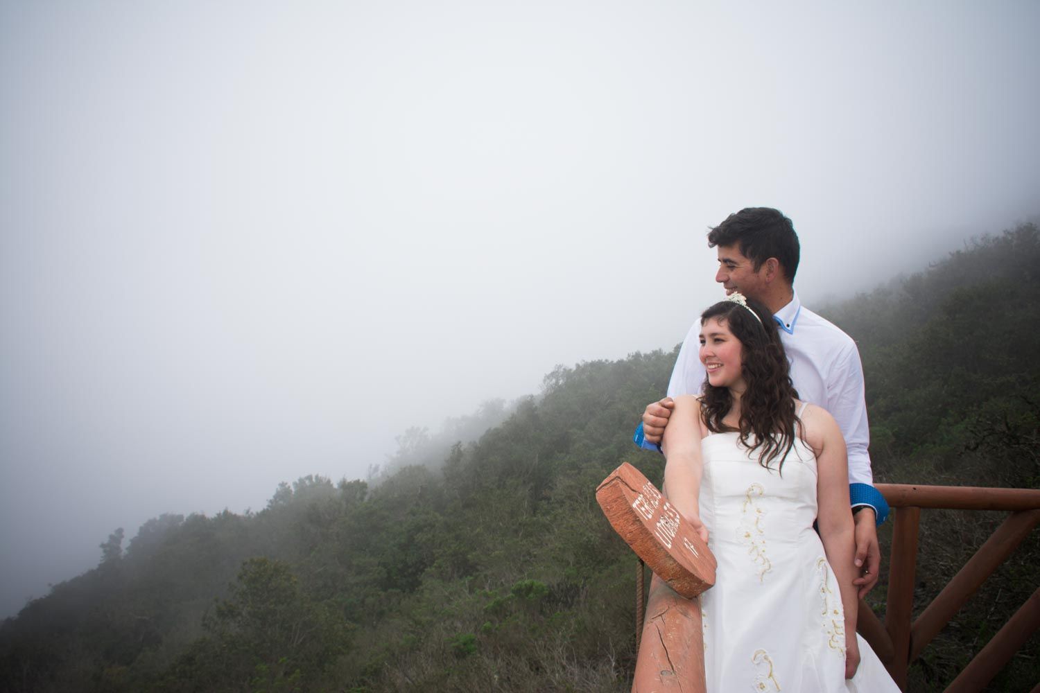 Bride and groom embrace on a misty overlook with foggy green hills in the background.