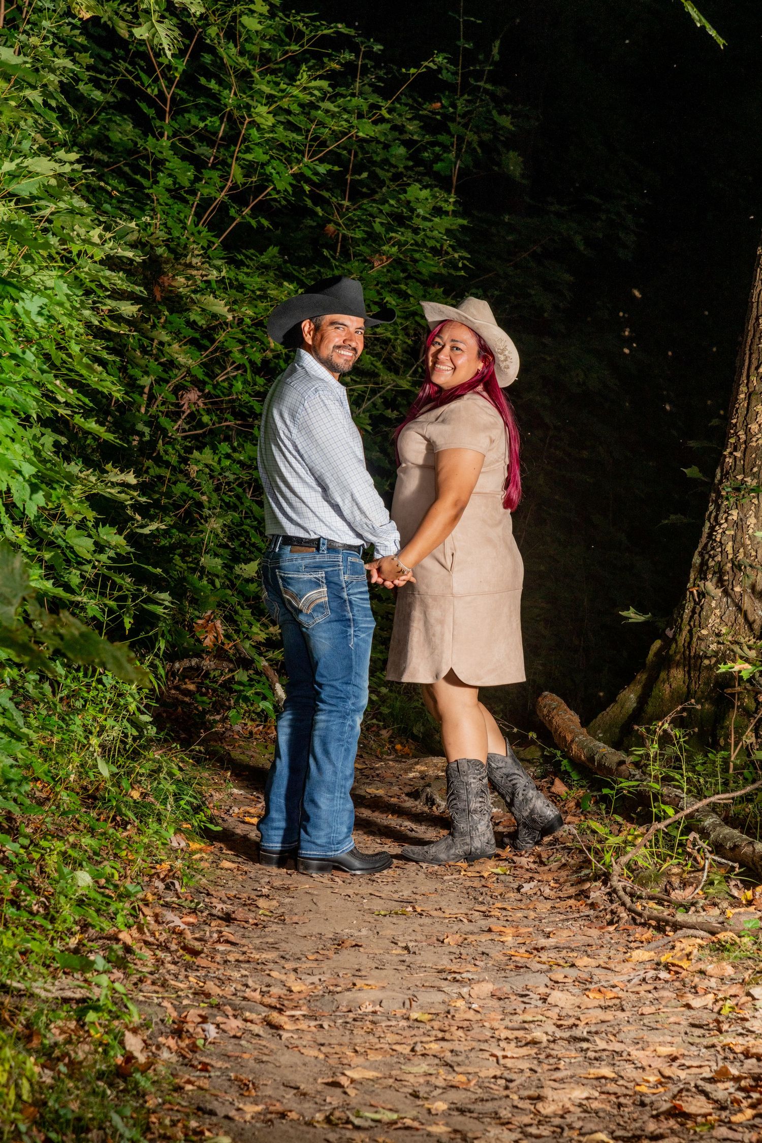 Happy couple in matching cowboy hats holding hands on a leaf-covered forest path at sunset.