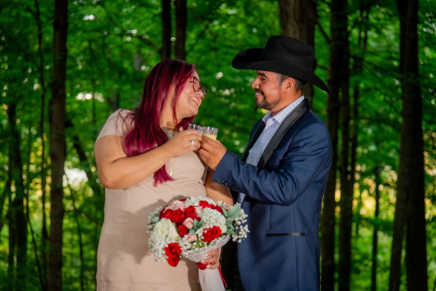Couple toasting with glasses in green forest, woman holding red and white bouquet wearing beige dress.